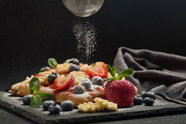 Waffles com frutas, hortelã e molho doce em tábua de servir em fundo escuro. Processo de polvilhar açúcar de confeiteiro sobre waffles.