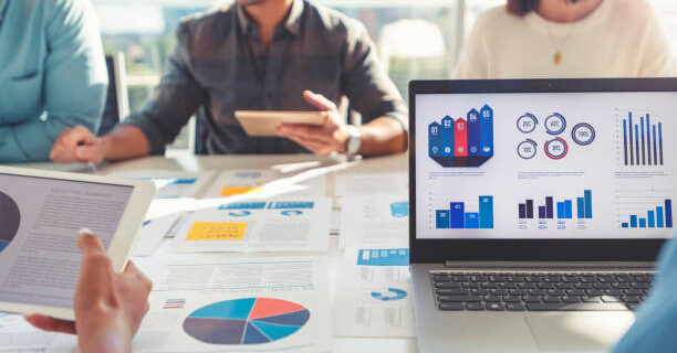 Group of people meeting with technology and paperwork. Close up of digital tablet and laptop computer with market analysis data on the screen. The documents have financial or marketing figures, graphs and charts on them. Multi ethnic group including Caucasian, latin and African American