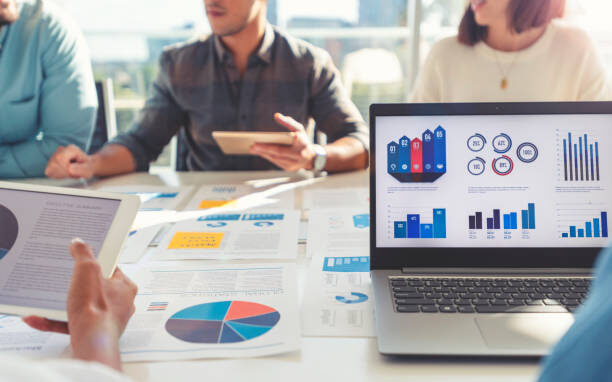 Group of people meeting with technology and paperwork. Close up of digital tablet and laptop computer with market analysis data on the screen. The documents have financial or marketing figures, graphs and charts on them. Multi ethnic group including Caucasian, latin and African American