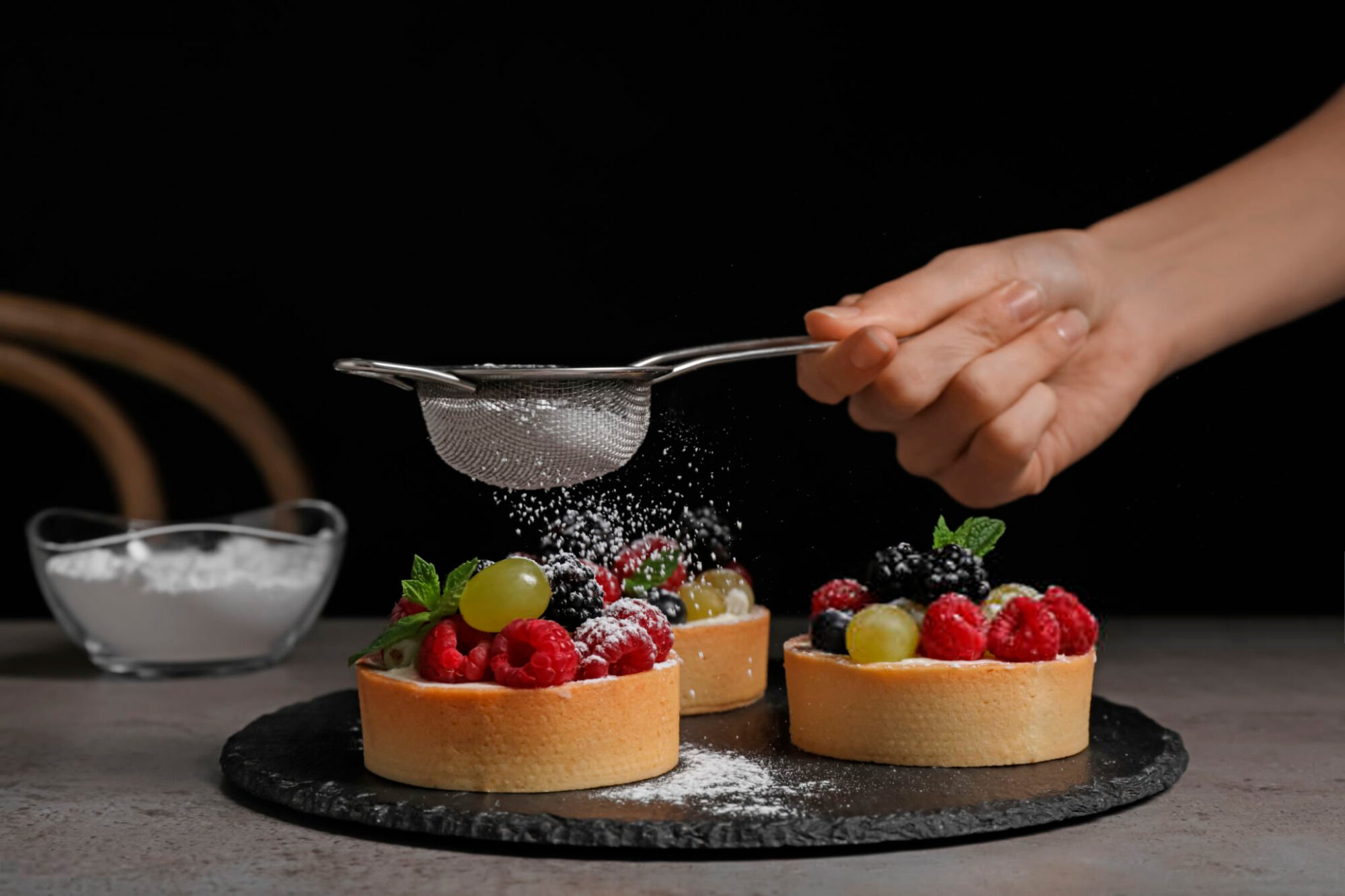 Woman sprinkling delicious tartlets with powdered sugar at grey table