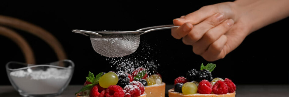 Woman sprinkling delicious tartlets with powdered sugar at grey table